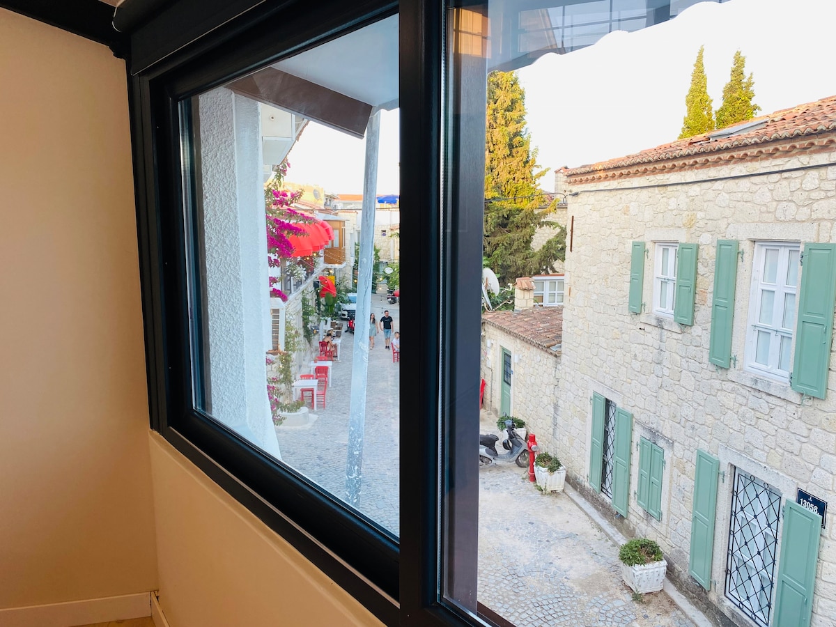 A view from a window frames a charming street lined with cobblestones. Colorful flowers cascade over balconies, blending with the stone buildings, each adorned with green shutters. The scene suggests a lively atmosphere, with seating areas and pedestrians visible along the walkways.