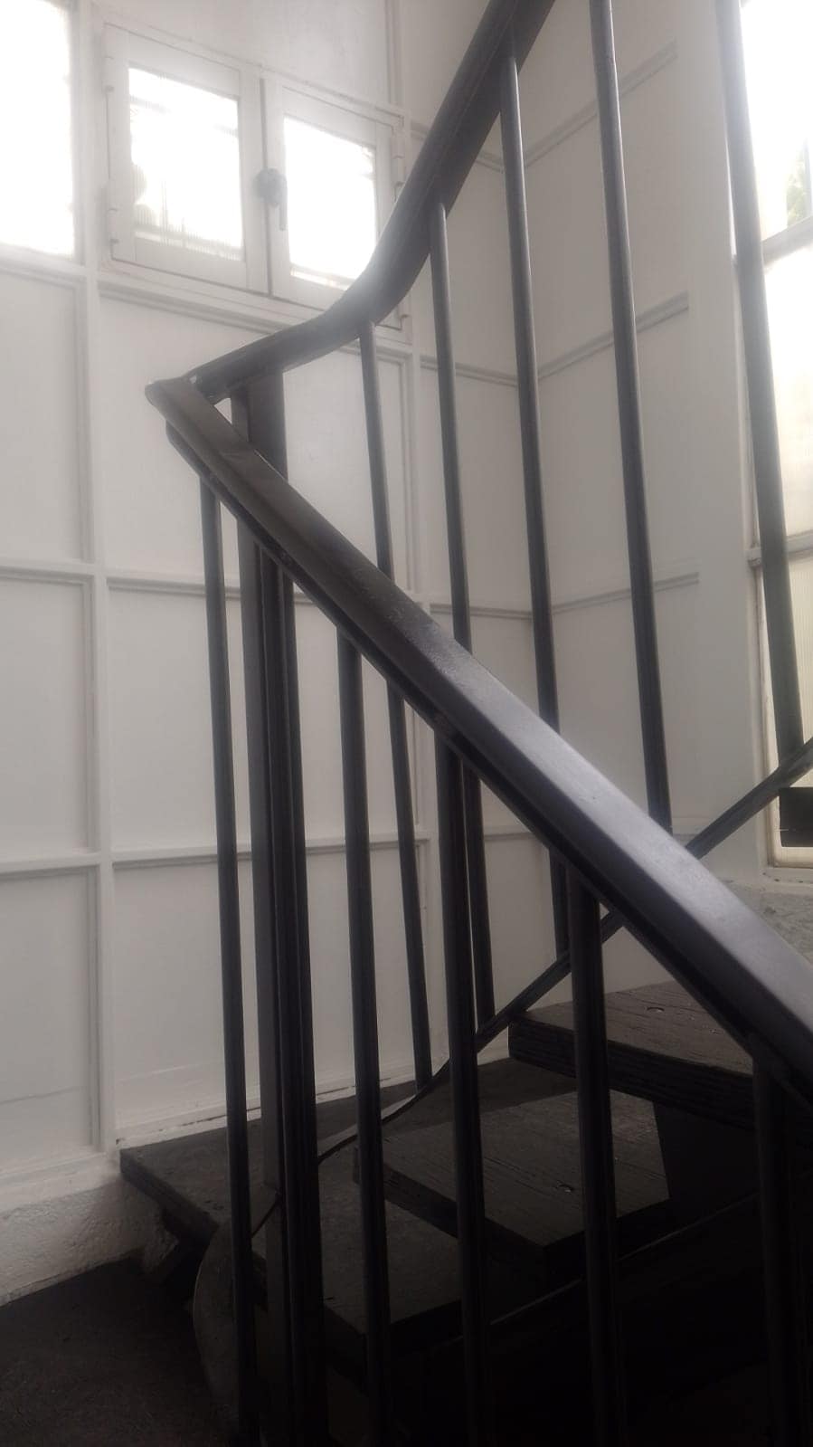 A staircase is shown with sleek black railings, leading upwards. The walls are painted white, and large windows allow natural light to filter in, creating a bright and open feel.
