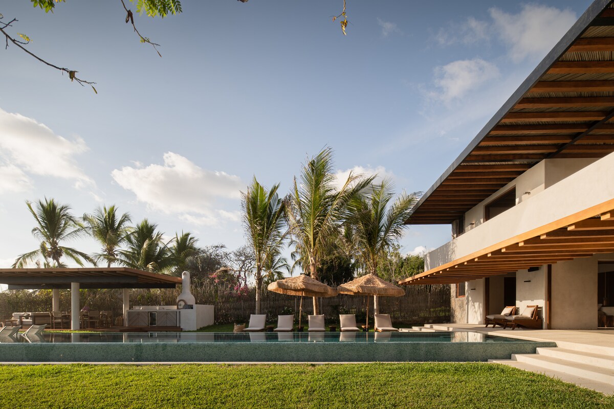 The outdoor area showcases a refreshing swimming pool, surrounded by lounge chairs and large umbrellas. A shaded patio area is present, featuring a sofa set. Lush green grass extends around the pool, with palm trees visible in the background against a clear sky.