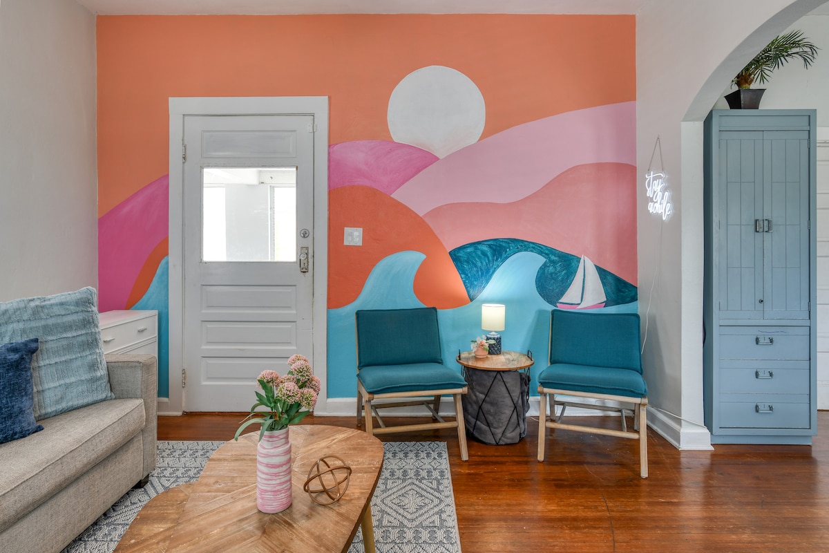A cozy living area features a vibrant mural depicting rolling hills and a sun setting over water. Two blue chairs are positioned beside a small table, which holds a lamp and a decorative item. A neutral sofa and a light-colored rug complete the inviting space.