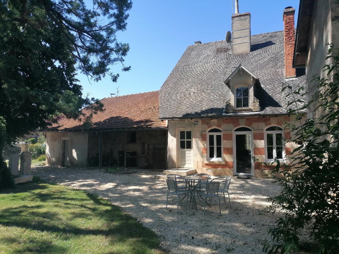 The exterior of a charming stone house is framed by lush greenery, showcasing a gravel area with a small table and chairs. The inviting entrance features windows and doors, while a textured slate roof complements the rustic design. Large trees provide shade to the surroundings.