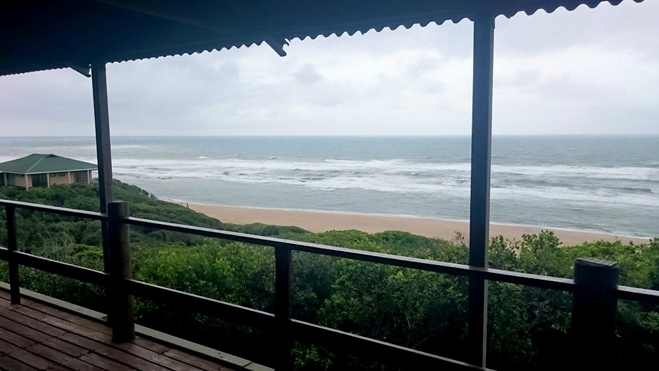 A wooden deck is showcased, offering a clear view of the sea and sandy beach. Gentle waves roll onto the shore, surrounded by greenery. Cloudy skies create a serene atmosphere, while the distant building is partially visible on the left.