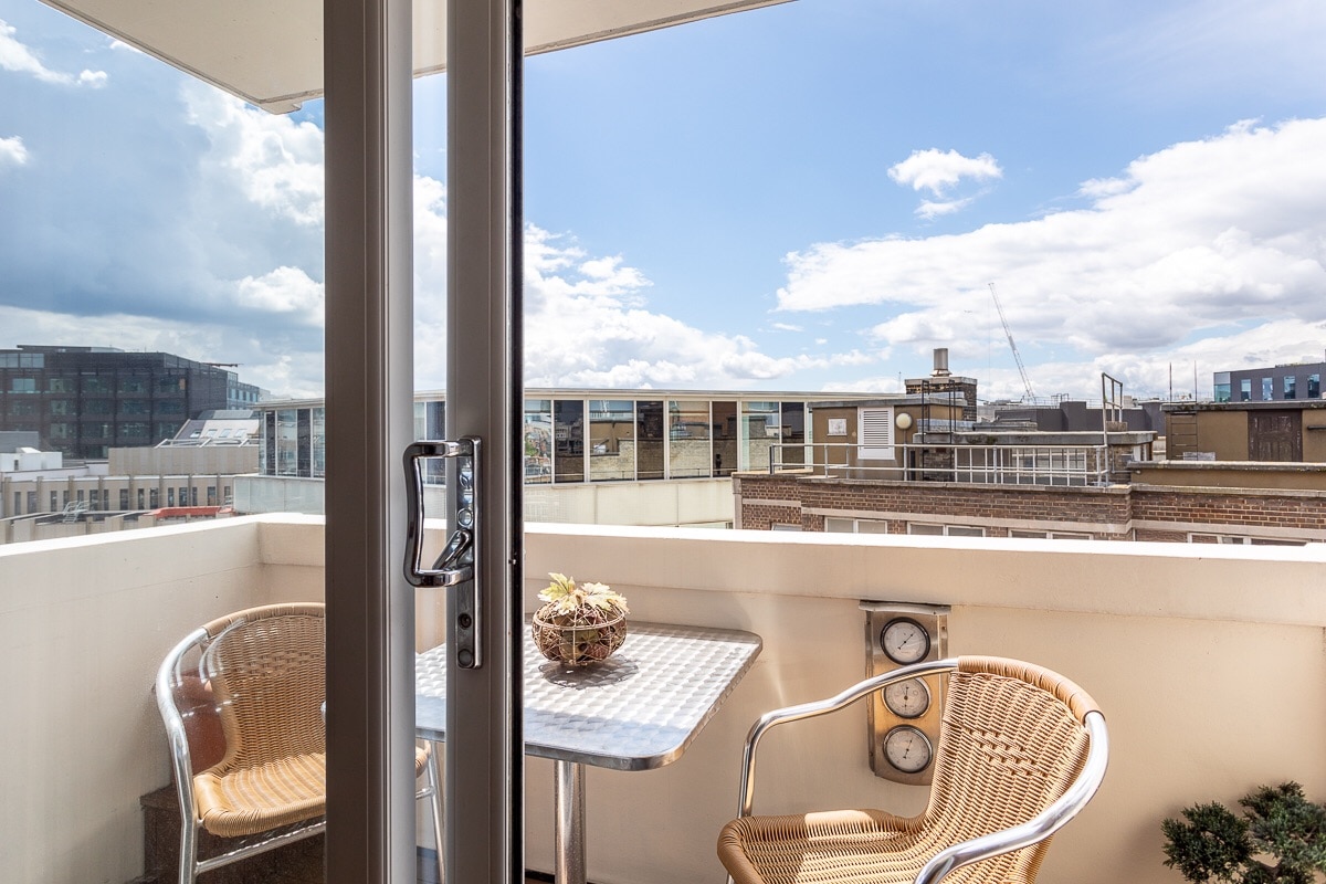 A small balcony is visible, featuring a round table and two wicker chairs. The view showcases a mixture of rooftops and buildings under a partly cloudy sky, with light filtering through the open door.