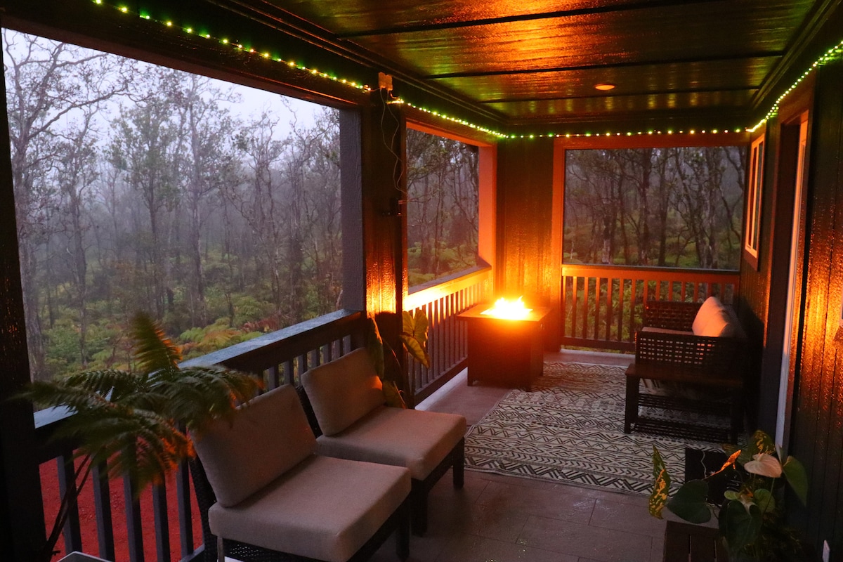 A cozy outdoor lanai features two cushioned chairs and a textured area rug, surrounded by lush greenery. Soft lighting lines the perimeter, creating a warm ambiance. Views of the misty ʻōhiʻa forest are visible beyond the railing, evoking a serene connection with nature.