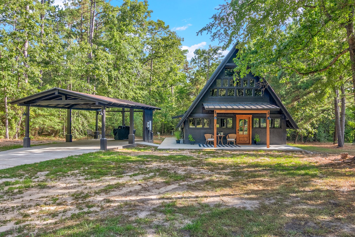An A-frame structure is surrounded by lush greenery, featuring large windows that allow natural light to enter. A covered parking area is visible next to the house, providing shaded space. Outdoor seating is arranged across from the entry, offering a welcoming spot to relax.