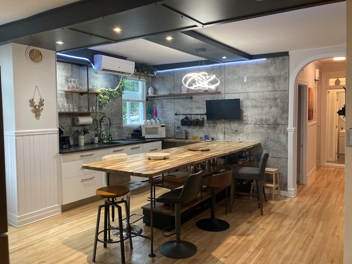 A modern kitchen area features a spacious wooden table surrounded by various seating options. Open shelves display glassware, while an air conditioning unit is mounted above the sink. A television is mounted on the wall, providing convenient entertainment. Natural light enters through a window.