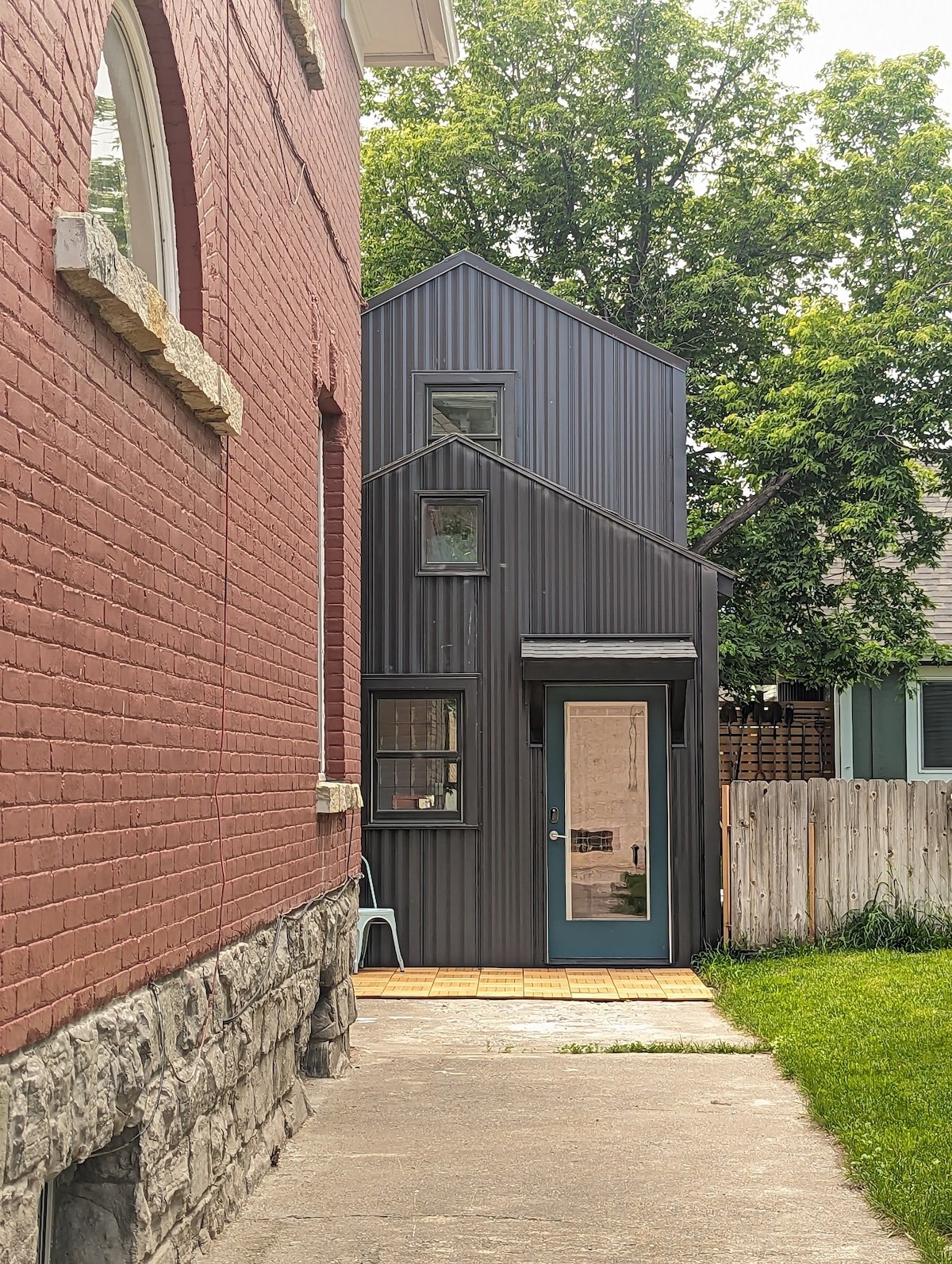 A narrow pathway is flanked by brick and stone walls, leading to a uniquely designed metal-clad structure. The building features large windows and a bright door, surrounded by grassy areas and trees, creating a connection to the outdoor environment.
