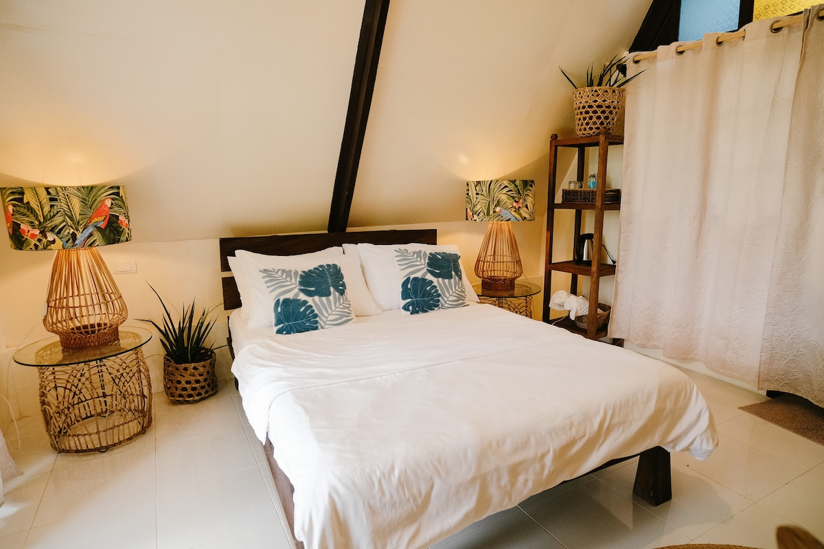 A cozy bedroom features a double bed with soft linens, flanked by bedside tables and woven lampshades. Natural textures and tropical accents adorn the space, complemented by decorative plants. A wooden shelf holds neatly arranged towels, enhancing the serene aesthetic of the room.