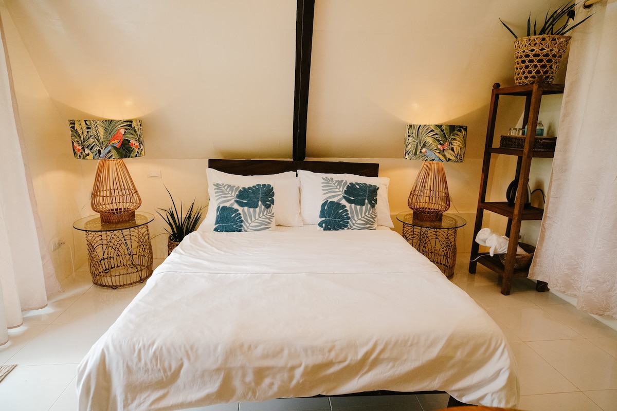 A cozy bedroom is furnished with a double-sized bed dressed in white linens. On either side of the bed, stylish wicker lampshades provide soft lighting. Tropical-themed decor enhances the ambiance, while a wooden shelf displays towels and plants, adding a touch of nature.