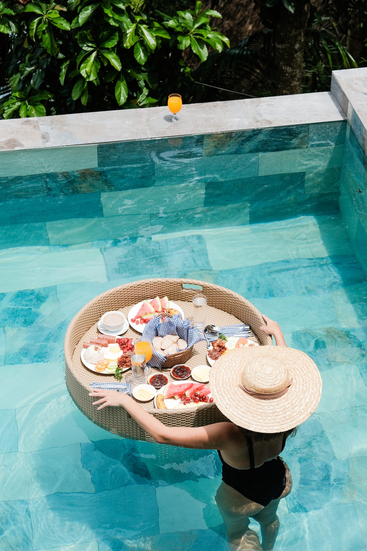 A woman stands in the infinity pool, holding a round floating tray filled with an assortment of breakfast items including fruits, pastries, and beverages. The tranquil water reflects the surrounding greenery, creating a serene atmosphere.