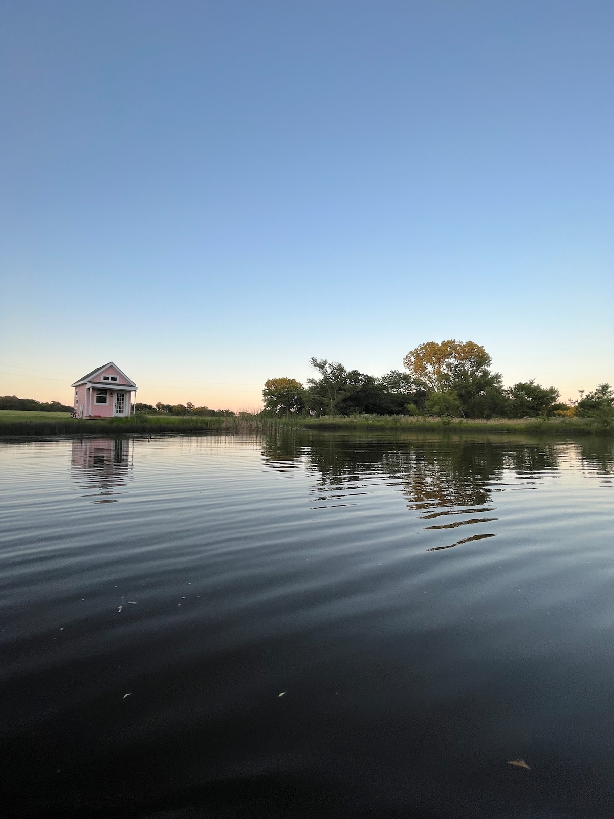 A charming pink cabin is reflected in the calm waters of the pond, surrounded by lush greenery and trees. The sky transitions from blue to warm hues near the horizon as dusk approaches, creating a serene atmosphere.