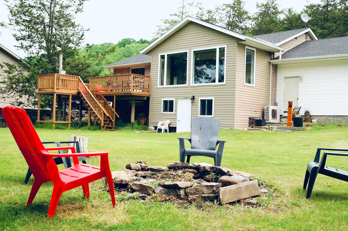 A spacious backyard features a fire pit surrounded by several outdoor chairs. The cottage with large windows is visible in the background, and a wooden deck extends from the home, blending seamlessly with the lush green lawn.