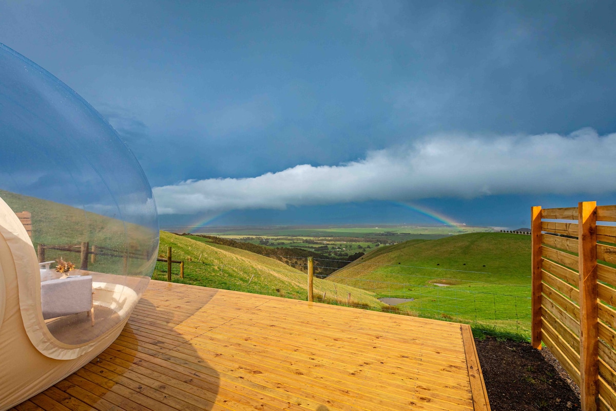 A transparent bubble structure is situated on a wooden deck overlooking rolling hills. A vibrant rainbow arcs across the cloudy sky, adding color to the serene landscape. The scene captures the tranquil connection between nature and modern comfort.