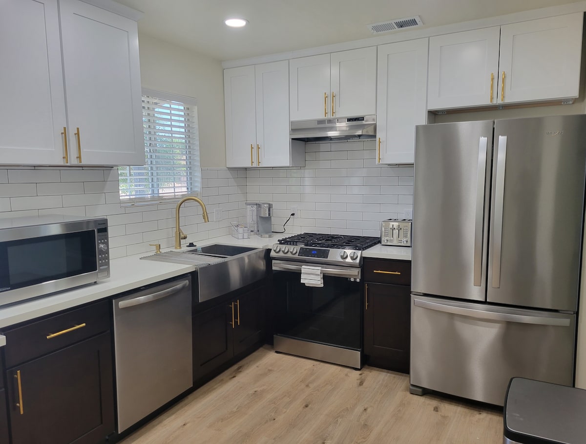 The kitchen features modern cabinetry with a mix of white and dark finishes, stainless steel appliances including a gas stove, refrigerator, and microwave. A double-basin sink with a gold faucet sits between the appliances. Light streams in through a window adorned with white blinds.