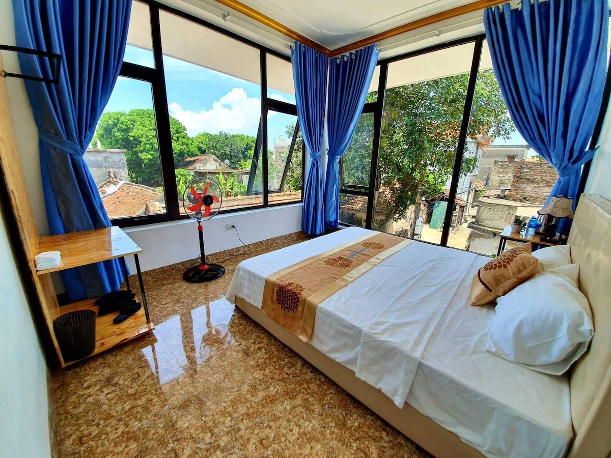 A bright bedroom features large windows adorned with flowing blue curtains, allowing natural light to fill the space. A spacious bed is positioned alongside a wooden shelf and a fan. Lush greenery is visible outside through the windows, providing a scenic view.