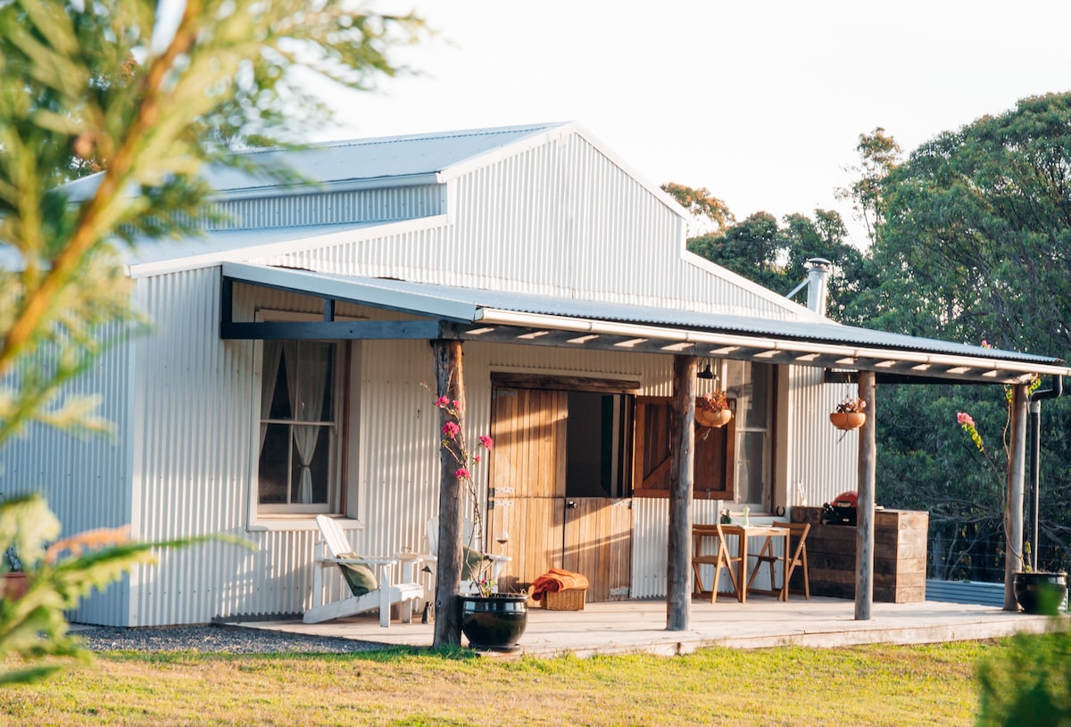 The eco cottage is shown from an angle that highlights its corrugated metal exterior and a spacious covered porch. Two white chairs and a small table adorn the patio, with hanging planters adding greenery. Large windows allow natural light to enter the inviting living space.