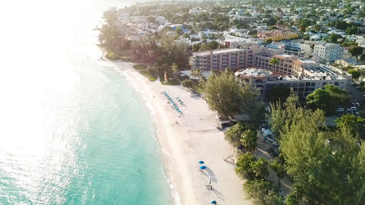 An aerial view reveals a serene beach lined with soft, white sand and gentle turquoise waves. Beach umbrellas are positioned along the shore, while lush greenery frames the coastal area. A low-rise building is visible in the background, complementing the inviting atmosphere of the seaside landscape.