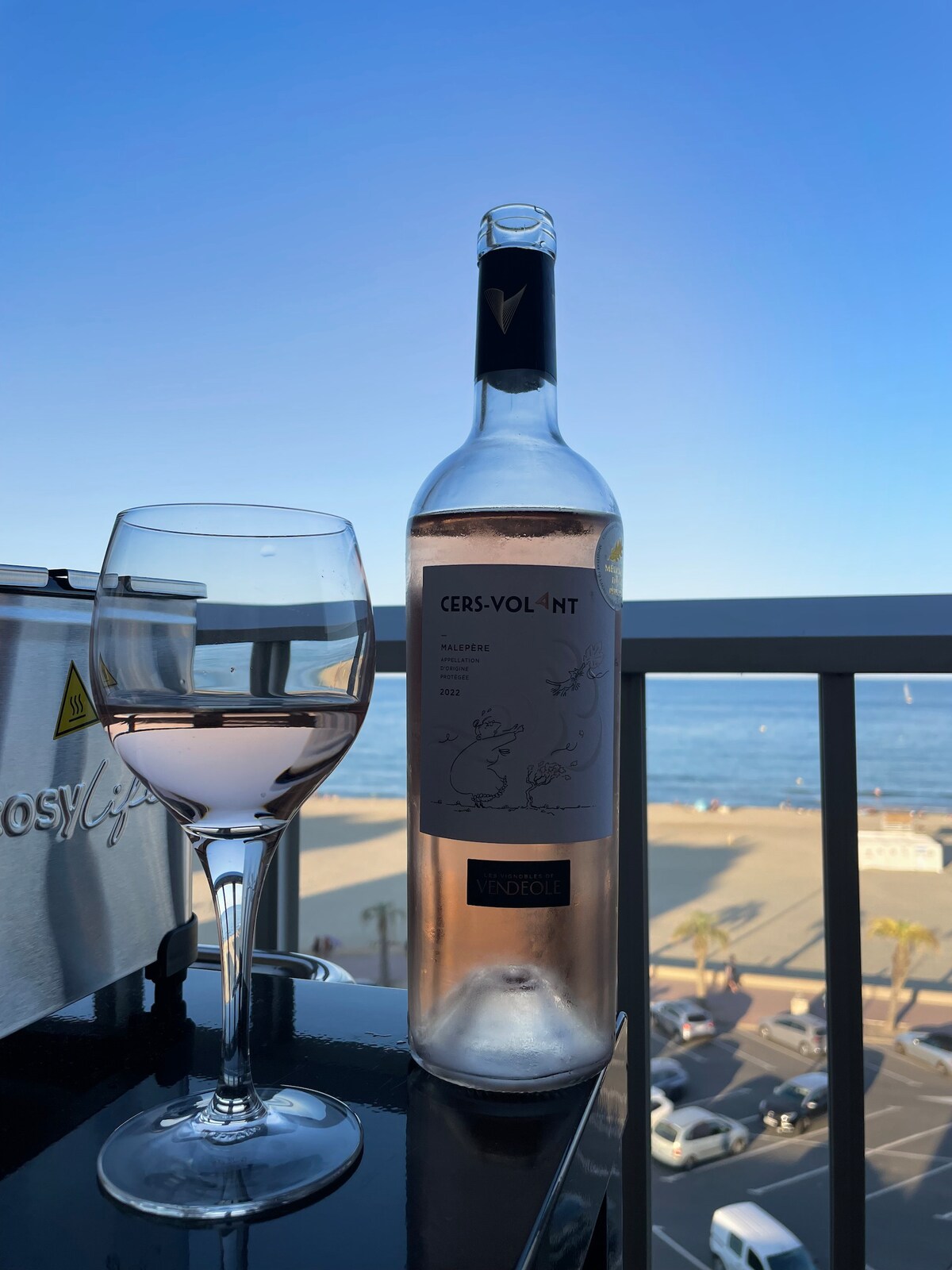 A bottle of rosé wine sits beside a glass filled with the same wine, placed on a balcony table. The background features a clear blue sky and a view of the ocean, with beach scenes and palm trees visible below.