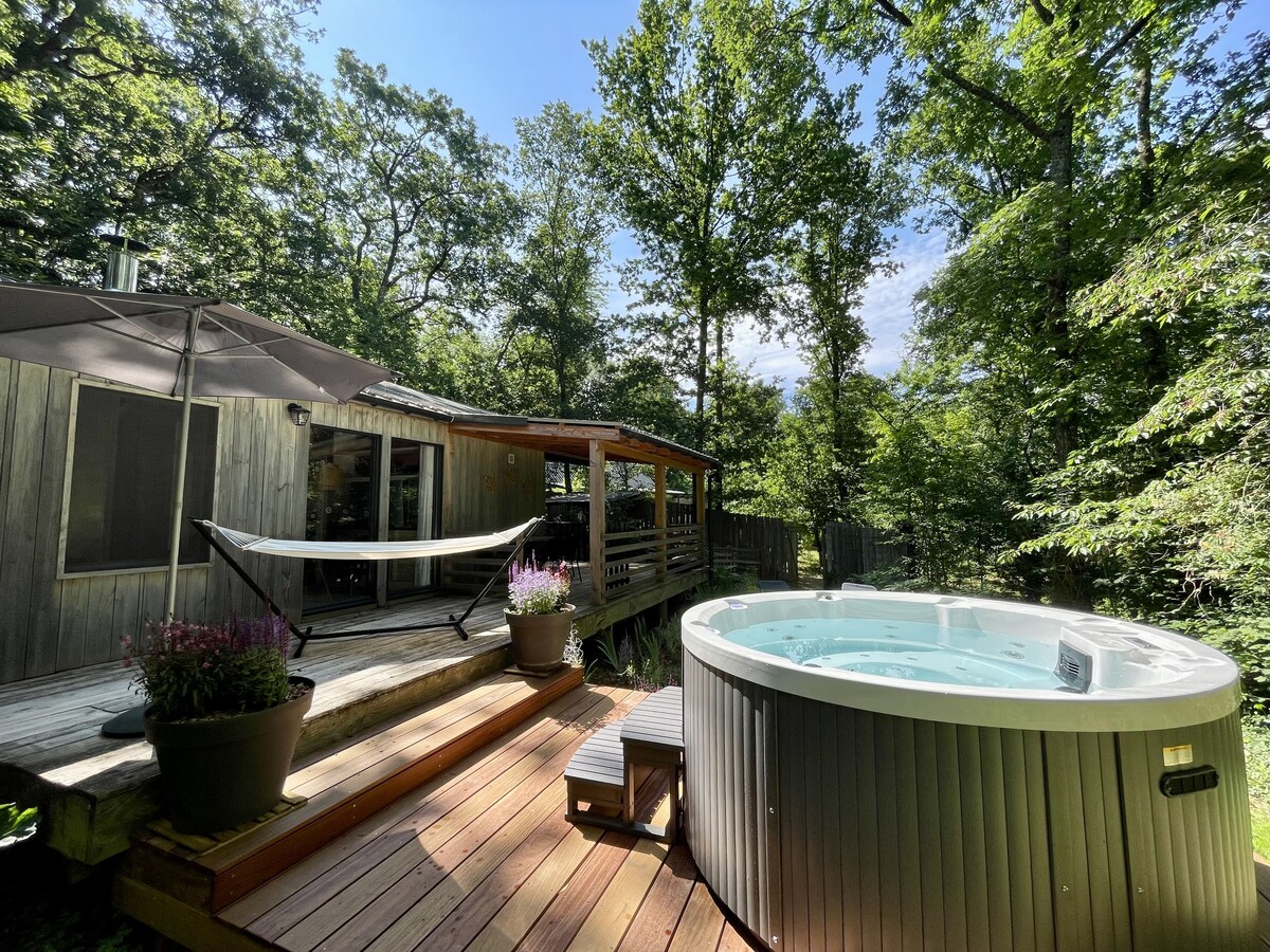 A private spa is positioned on a wooden deck surrounded by lush greenery. A shaded seating area features a hammock and an umbrella, inviting relaxation. The cozy lodge is visible in the background, enhancing the serene outdoor atmosphere.