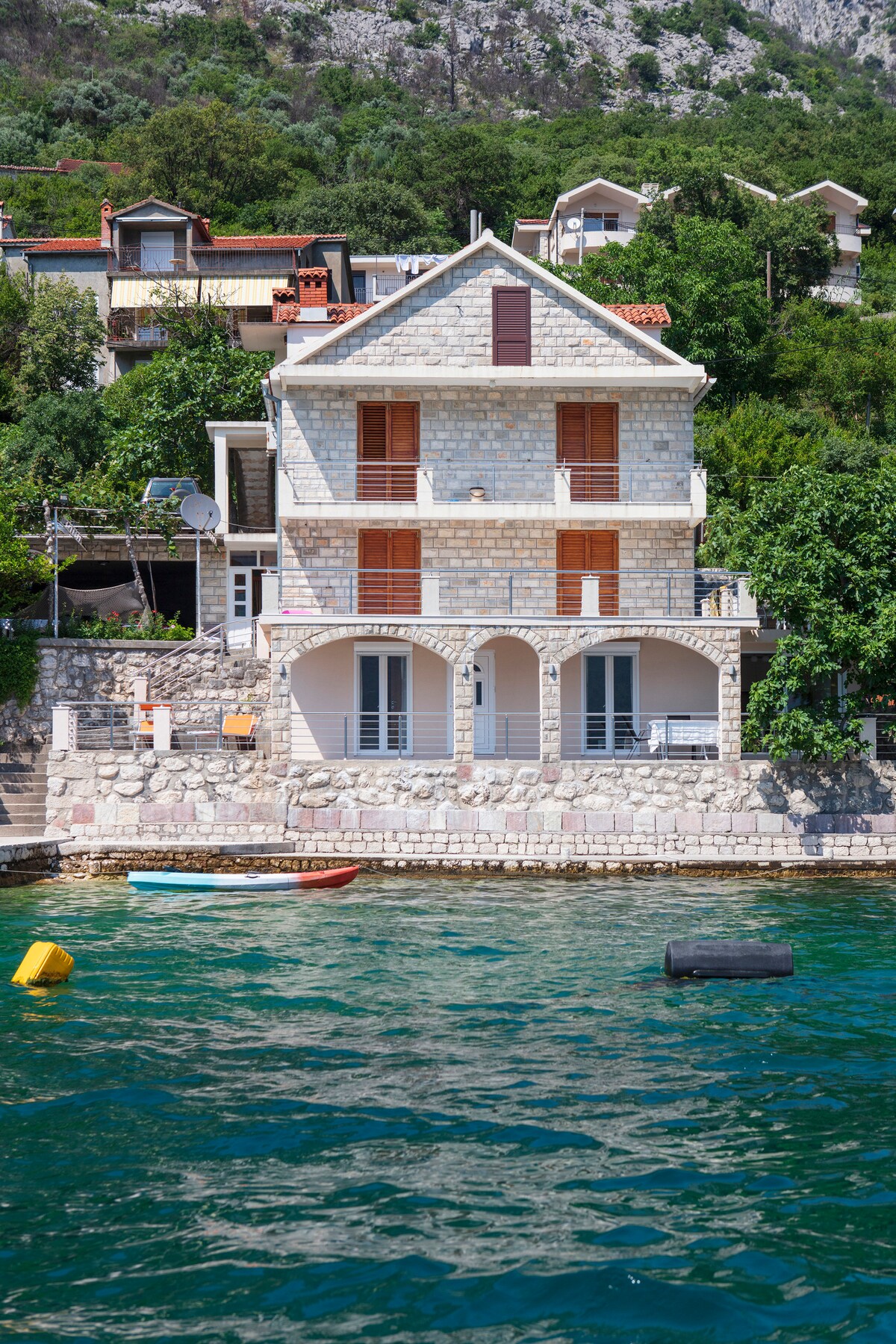 A two-story stone house features a classic architectural style, with wooden shutters on its windows. The structure is situated by the water's edge, surrounded by greenery, offering a peaceful view of the nearby boats floating in the clear water.