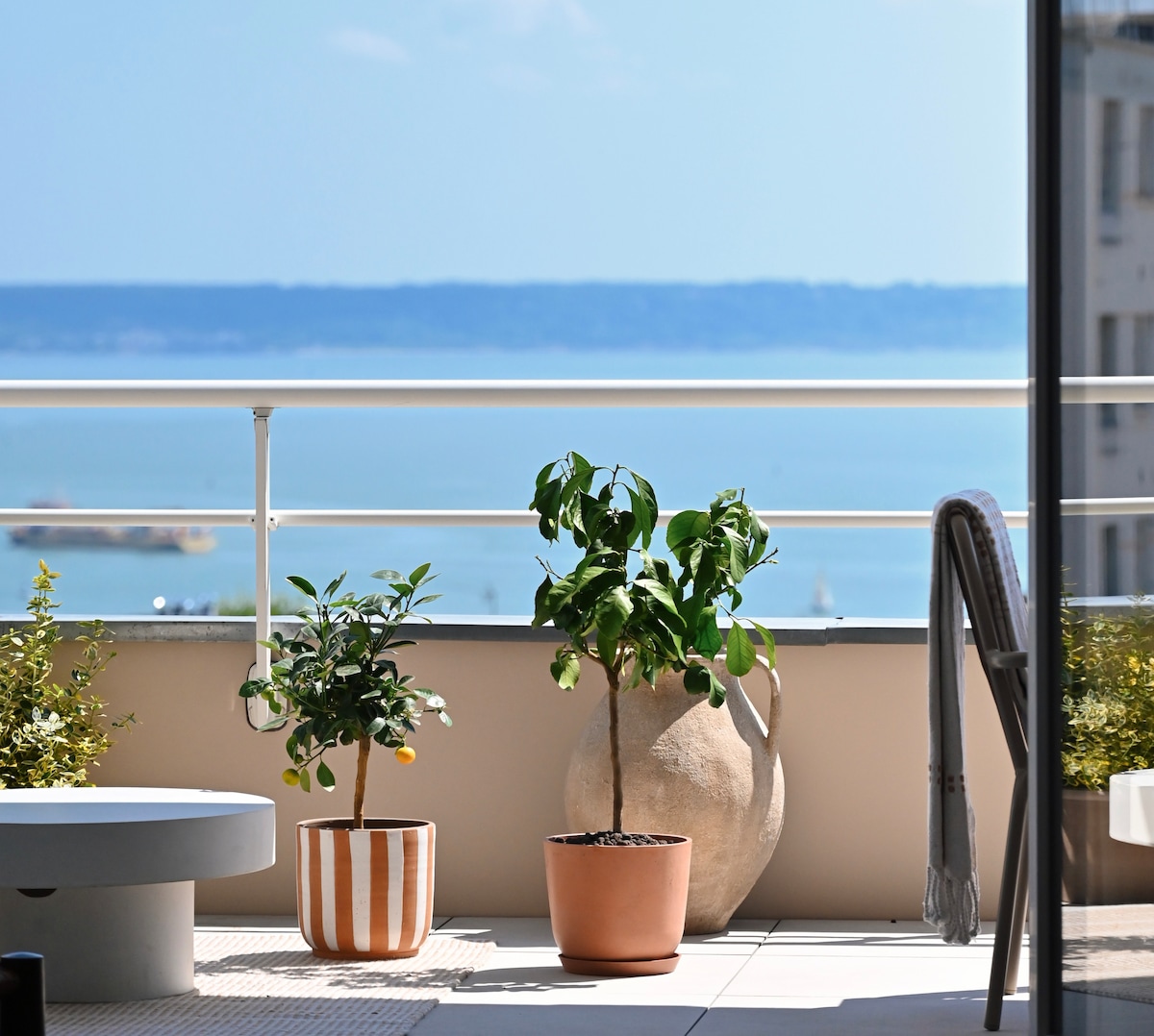 A terrace is shown with two potted plants, one with vibrant green leaves and the other featuring small citrus fruits. A smooth stone pot adds texture, while the sea view is visible in the background, creating a serene coastal ambiance.