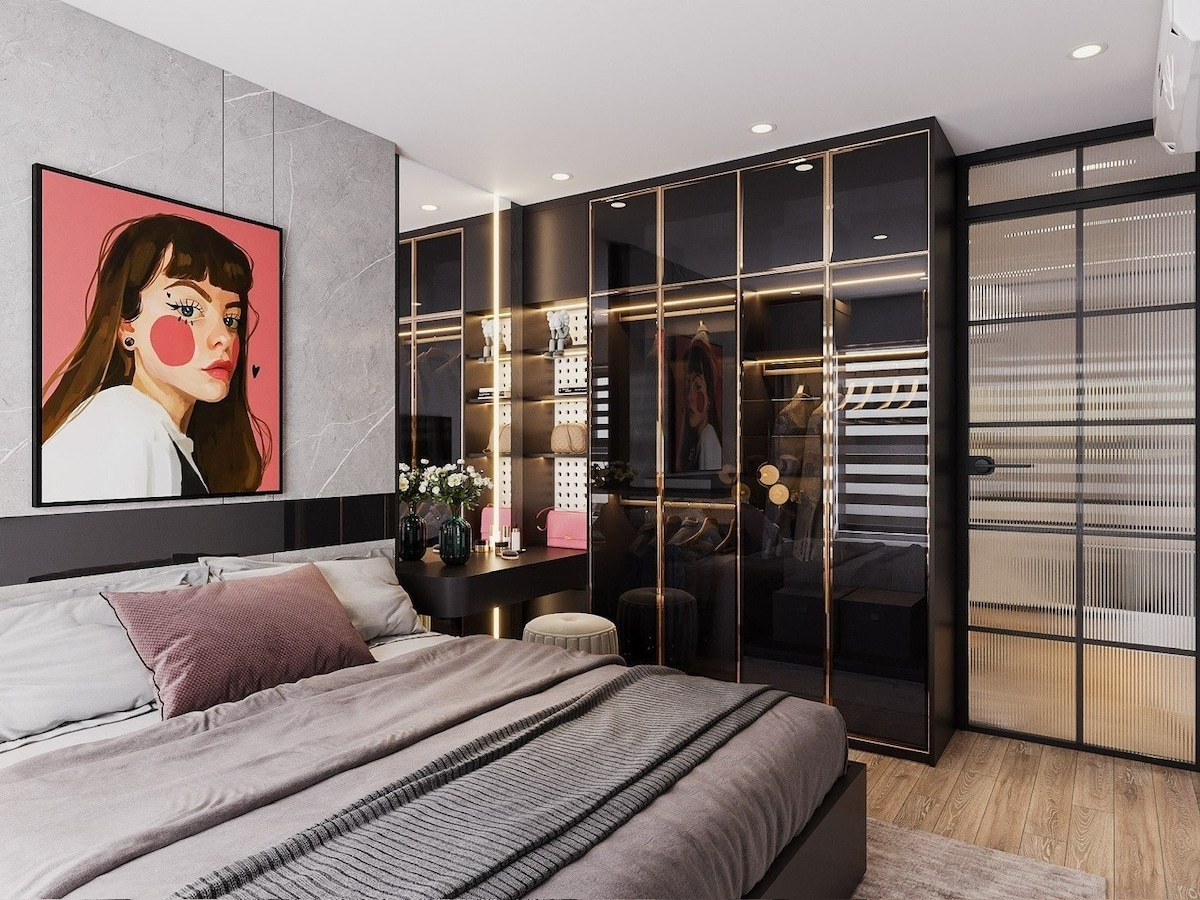A modern bedroom features a spacious bed covered with textured linens in neutral tones. A stylish wardrobe with glass doors is visible, alongside a sleek desk area adorned with flowers. An art piece hangs above the bed, complementing the sophisticated decor and lighting.