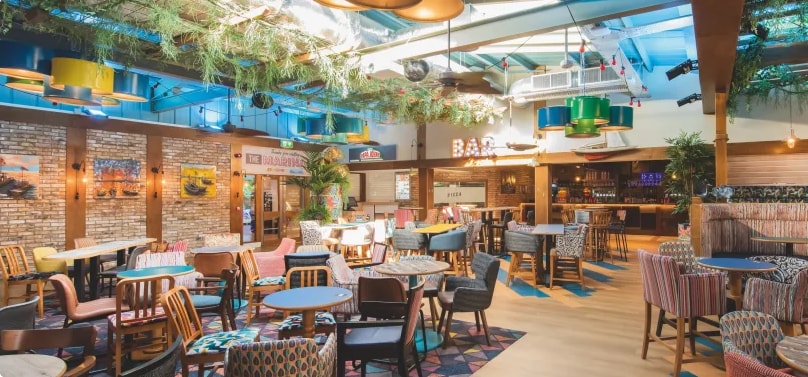 A spacious dining area is presented with a variety of colorful seating arrangements, including wooden and upholstered chairs. Decorative plants and overhead lights enhance the vibrant atmosphere, while tables are set up for both small and larger gatherings.