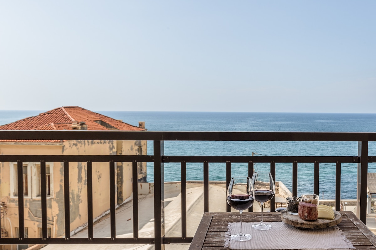 A balcony table is set with two glasses of red wine beside a plate of snacks. The view reveals an expansive sea and a glimpse of neighboring buildings, contributing to a serene atmosphere.