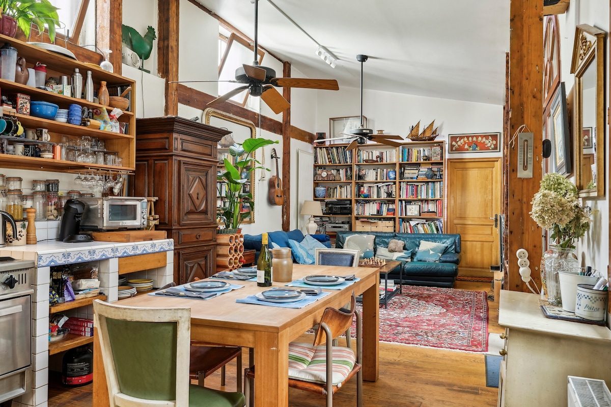 A spacious kitchen and living area features wooden beams and large windows that invite natural light. A dining table with place settings sits in the foreground, while a cozy seating area with blue sofas and a bookshelf filled with books is visible in the background.