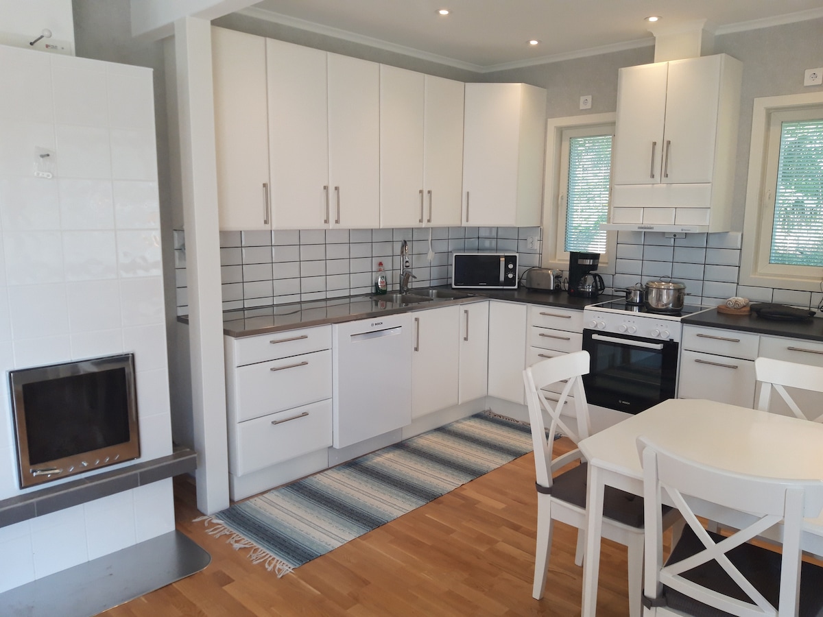 A modern kitchen features sleek white cabinetry and a spacious countertop. Appliances include a microwave, oven, and dishwasher. A dining table with four white chairs is positioned nearby, complemented by a rug in soft blue and beige tones. Natural light enters through multiple windows.