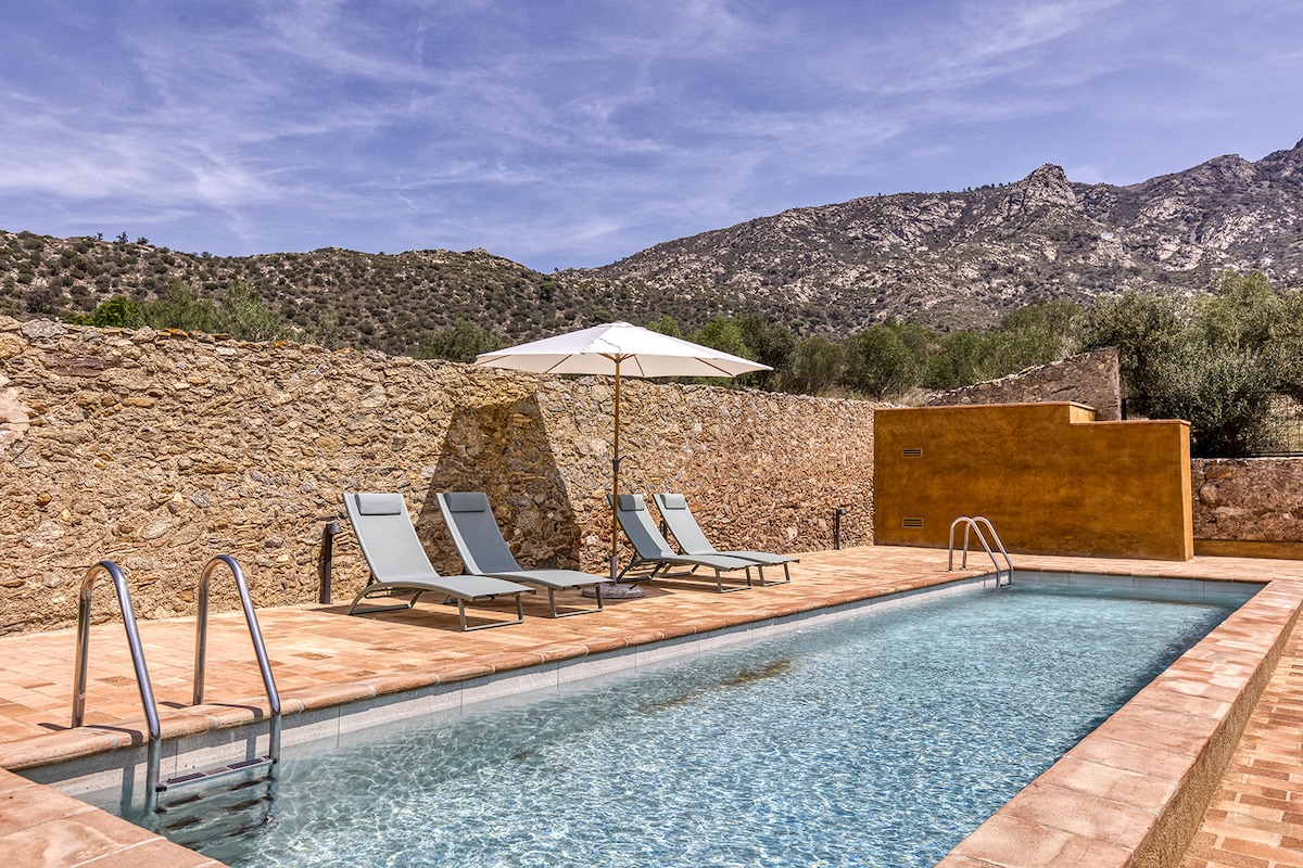 An outdoor pool is featured within a stone courtyard, complemented by two lounge chairs and a large umbrella. Surrounding mountains are visible in the background, providing a natural setting, while the stone wall offers privacy to the area.