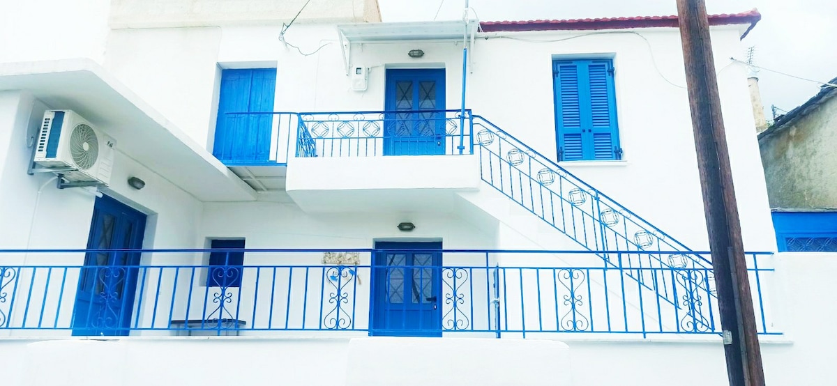 A traditional building showcases bright blue doors and railings against a crisp white exterior. The structure features a staircase leading to the upper level, with a decorative railing complementing the design. An air conditioning unit is positioned on the left side.