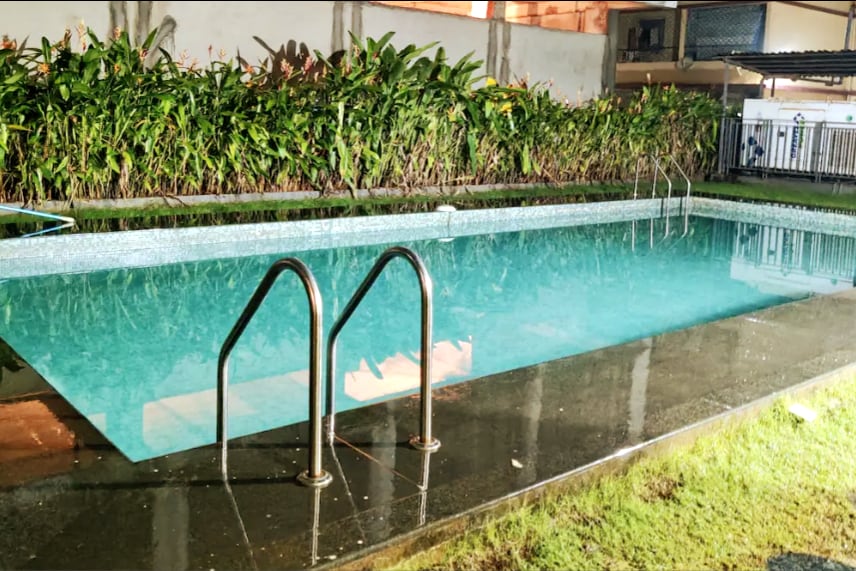 A shared swimming pool is visible, featuring a clear, turquoise surface. The pool is bordered by a sleek stone deck and surrounded by lush greenery. Two stainless steel handrails provide access to the water, illuminated softly by surrounding lights.