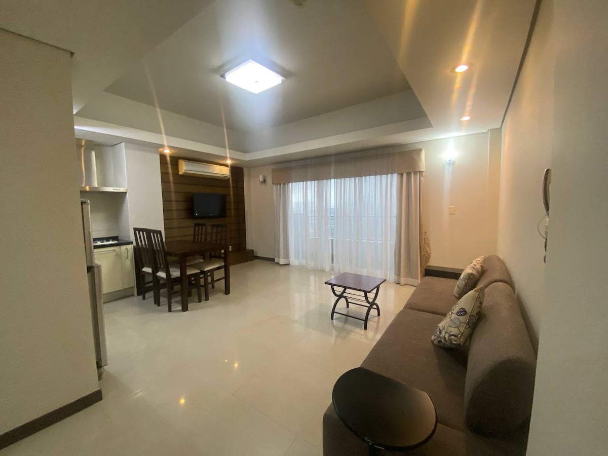 An open living area features a comfortable sofa paired with a small coffee table. In the background, a dining table with four chairs sits adjacent to the kitchen, while large windows allow natural light to brighten the space.
