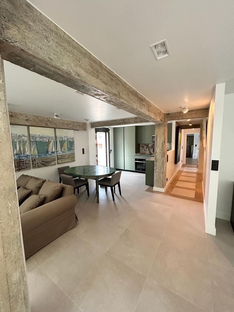 A modern living space features a spacious dining area with a glass table surrounded by four chairs. An open design connects to a well-equipped kitchen, with textured concrete beams adding structural interest. Natural light illuminates the room from a large window, enhancing the contemporary aesthetic.