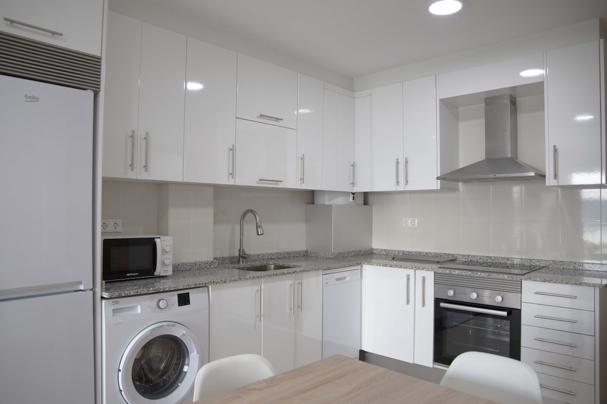 A modern kitchen is presented, featuring glossy white cabinets, a stainless steel stove, and a hood. A microwave and washing machine are visible, along with a composite countertop. A dining table with light-colored chairs is positioned in the foreground, providing a functional and inviting cooking space.