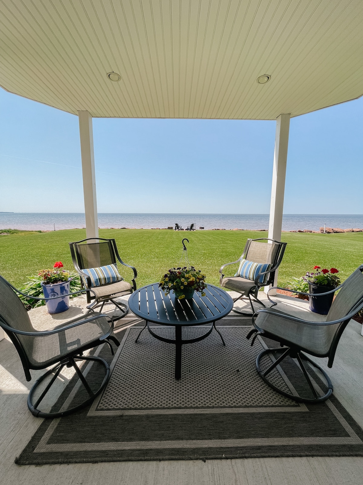 A covered patio area is furnished with four swivel chairs around a circular table. Potted flowers are placed around the space, and a scenic view of the beach and water is visible in the background beneath a clear sky.