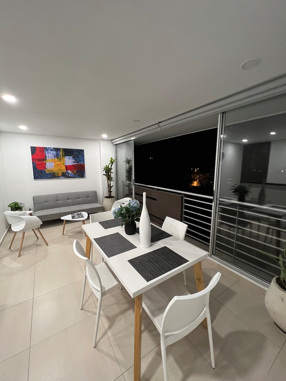 A modern dining area features a white table surrounded by four chairs. A gray sofa is positioned nearby, complemented by vibrant artwork on the wall. Large glass doors provide access to a balcony with views of the evening city lights.