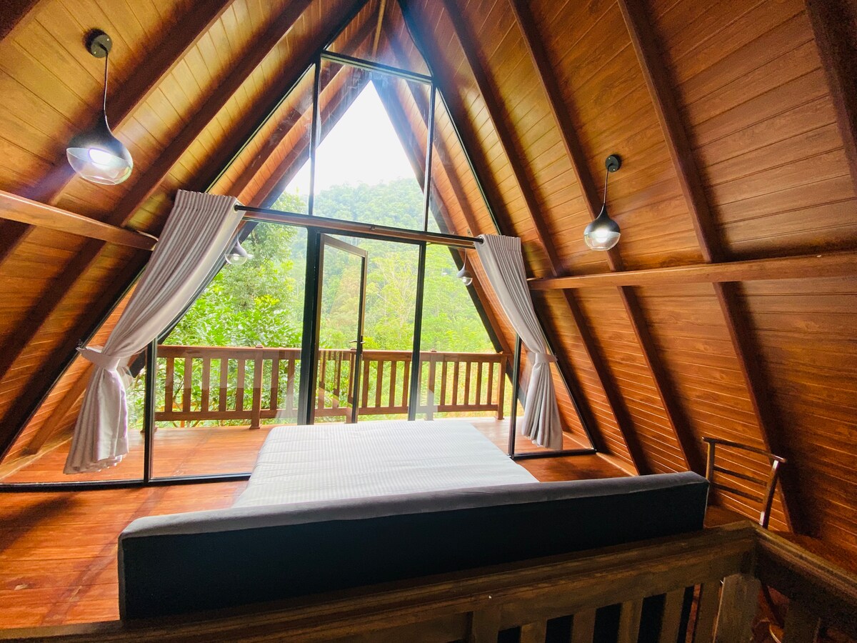 A spacious bedroom features wood-paneled walls and an A-frame ceiling, with large windows offering views of the surrounding greenery. Light-filtering curtains frame the windows, and a neatly made bed is positioned centrally, inviting relaxation and connection with nature.