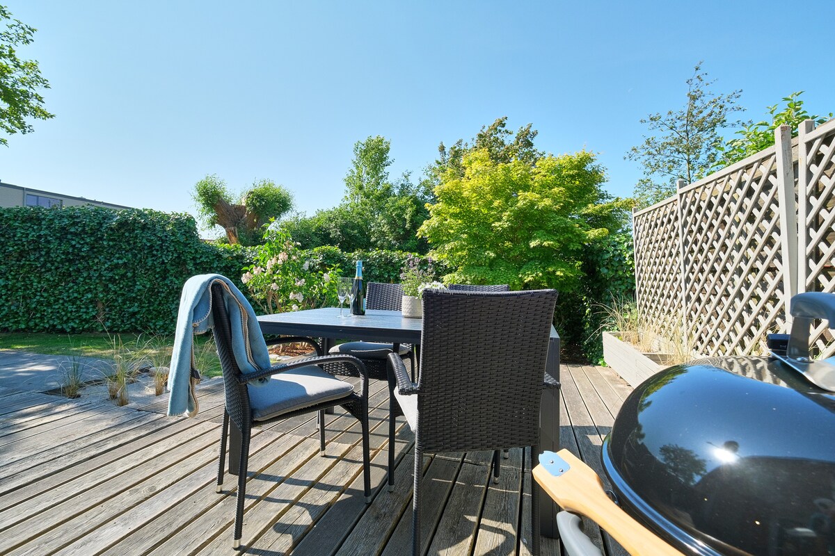 A spacious outdoor terrace is depicted, featuring a dining table surrounded by six wicker chairs. A black grill is positioned nearby, with greenery and a wooden privacy screen visible in the background, creating a serene setting for outdoor meals.