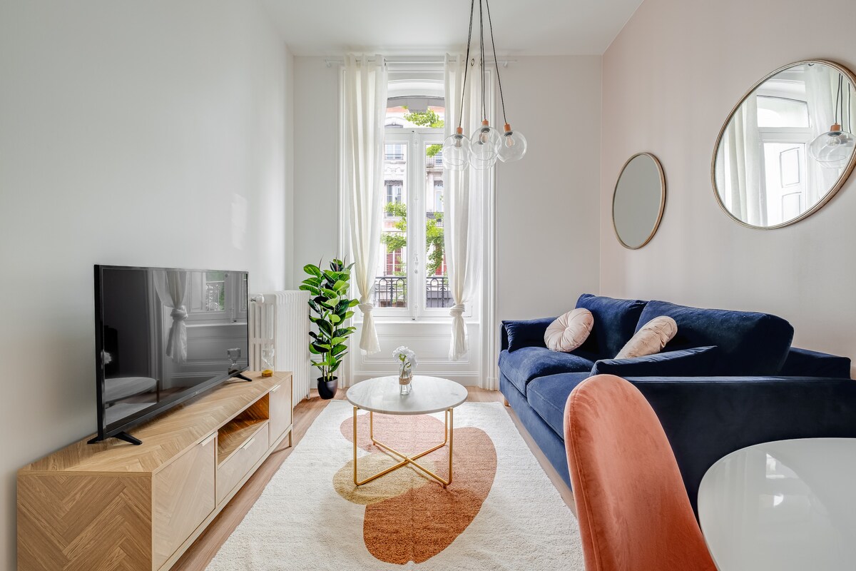 A cozy living area is presented, featuring a blue sofa and a round coffee table atop a soft area rug. A television is placed on a wooden media console. Natural light streams through large windows adorned with sheer drapes, illuminating a green plant in the corner.