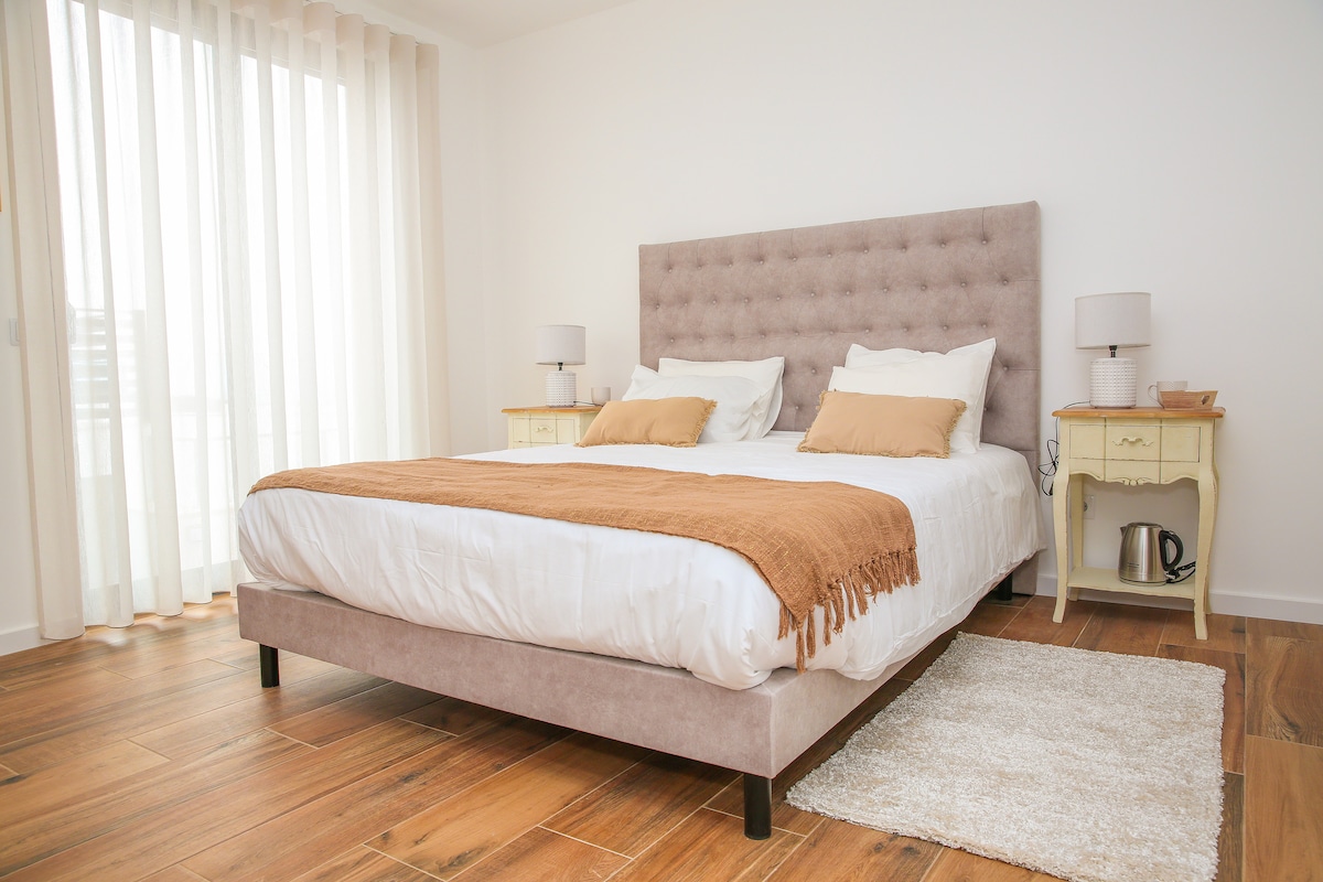 A spacious sleeping area features a king-size bed with a tufted headboard, dressed in neutral linens and accented with decorative pillows. Bedside tables with lamps flank the bed. Large sheer curtains allow natural light to fill the room, and wooden flooring adds warmth.