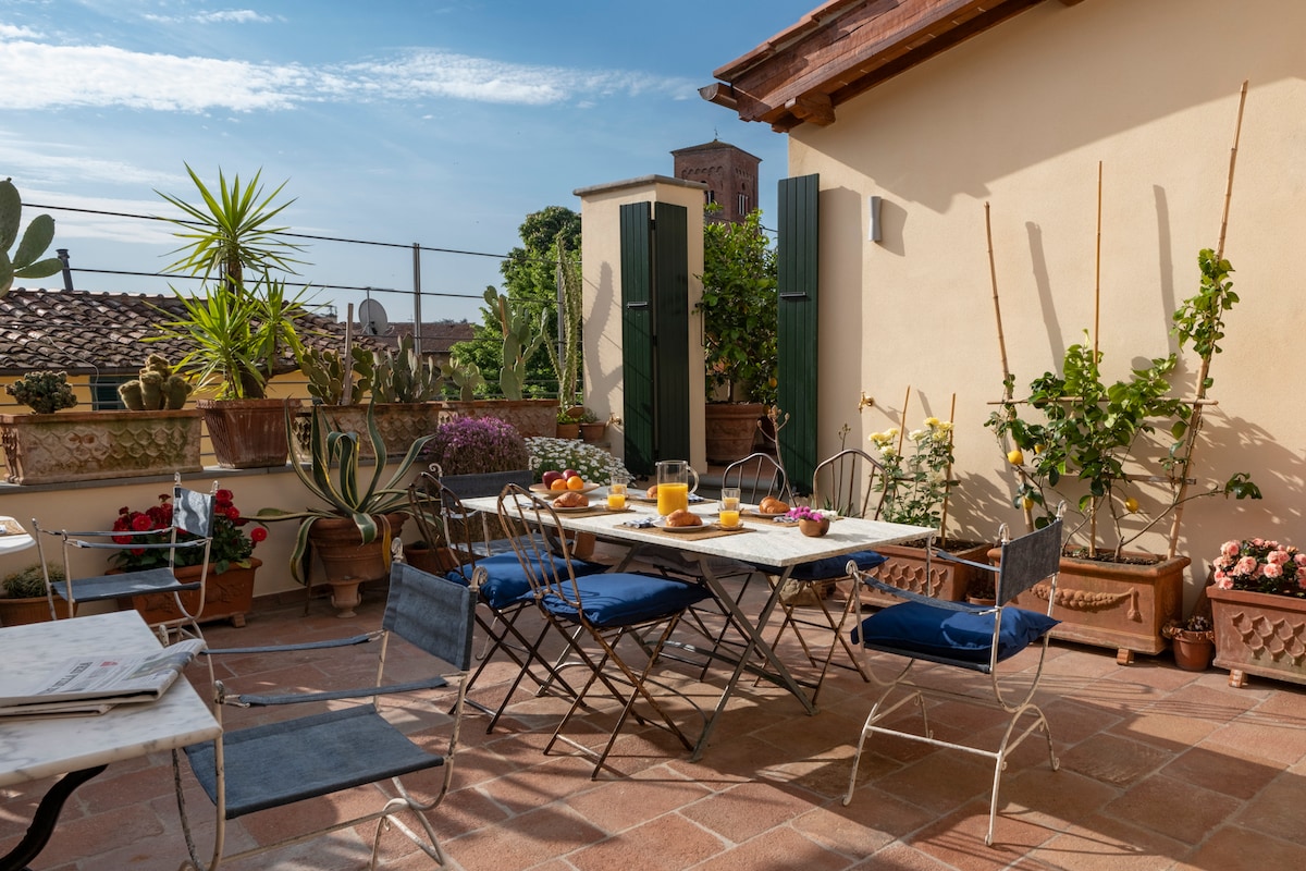 A spacious terrace is equipped with several tables and chairs, surrounded by potted plants and greenery. The table is set with glasses and pitchers, suggesting a welcoming space for breakfast or afternoon gatherings. Bright sunlight enhances the inviting ambiance.