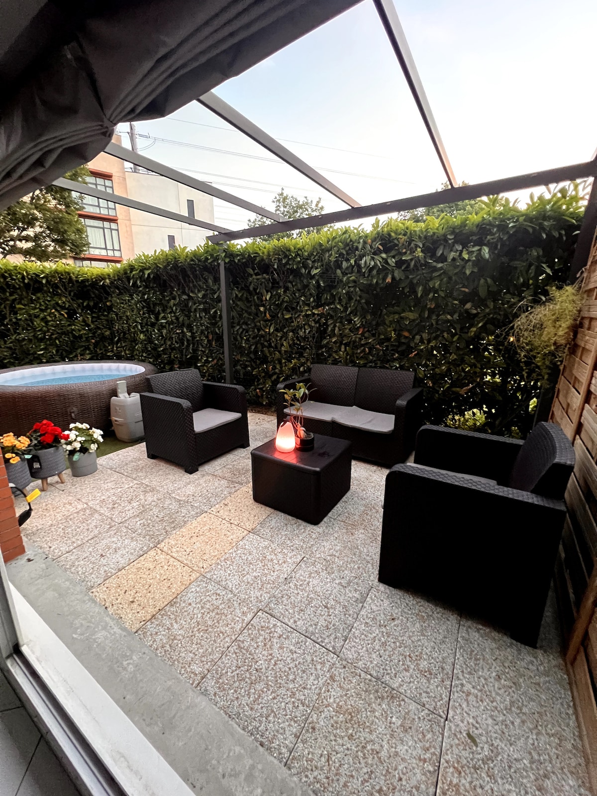 A cozy outdoor seating area is showcased, featuring a set of comfortable black chairs and a matching couch surrounding a central table. Verdant hedges provide privacy, and decorative plants add a touch of greenery. A spa can be seen in the background, enhancing the relaxation space.