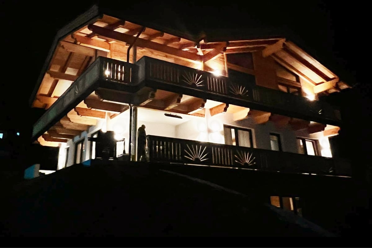 The exterior of the accommodation is illuminated at night, showcasing a charming wooden structure with a sloped roof and multiple balconies. Soft lighting highlights the architectural details, creating a welcoming presence. The surrounding area is dark, emphasizing the warm glow of the building.