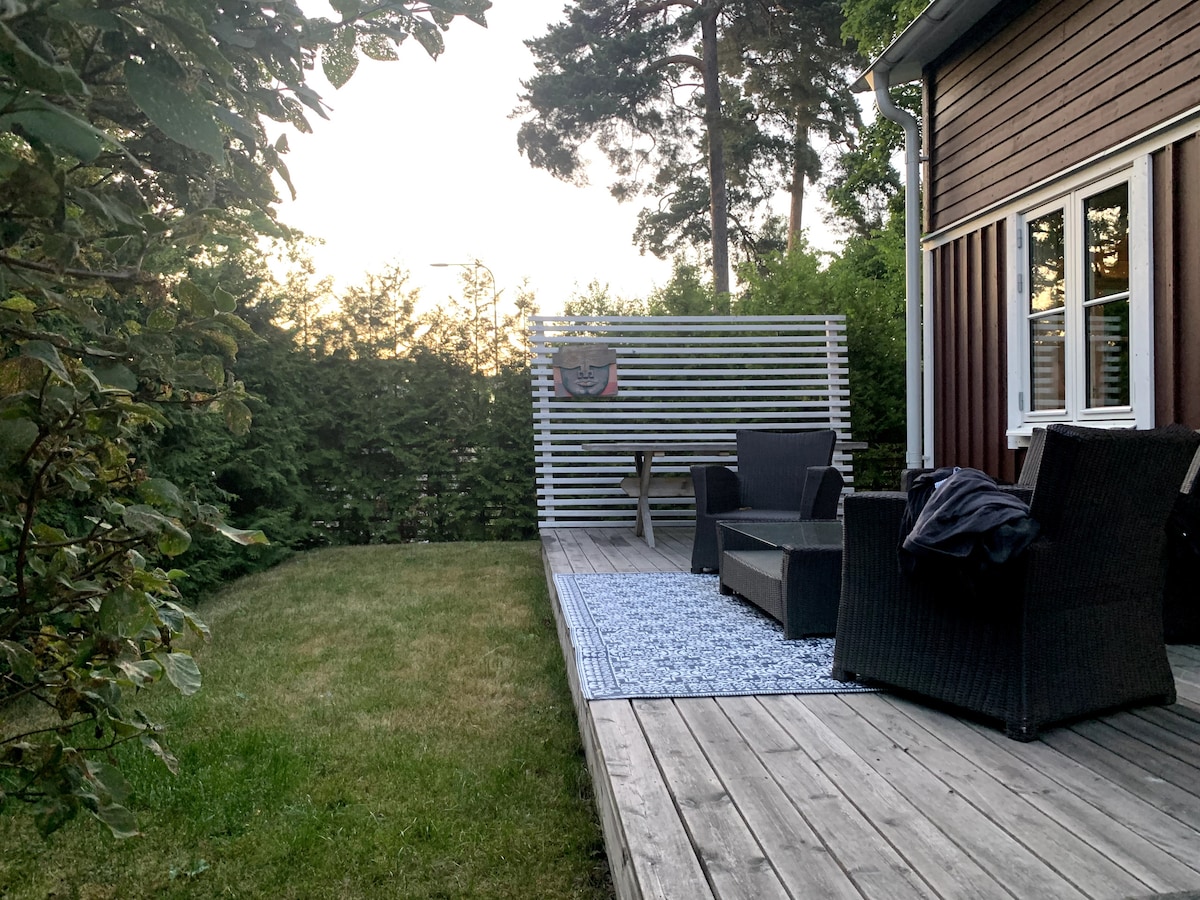 A wooden deck is featured, extending into a grassy backyard, framed by tall greenery for privacy. Two dark wicker chairs and a small table are positioned on the deck, while a patterned rug adds texture to the space.