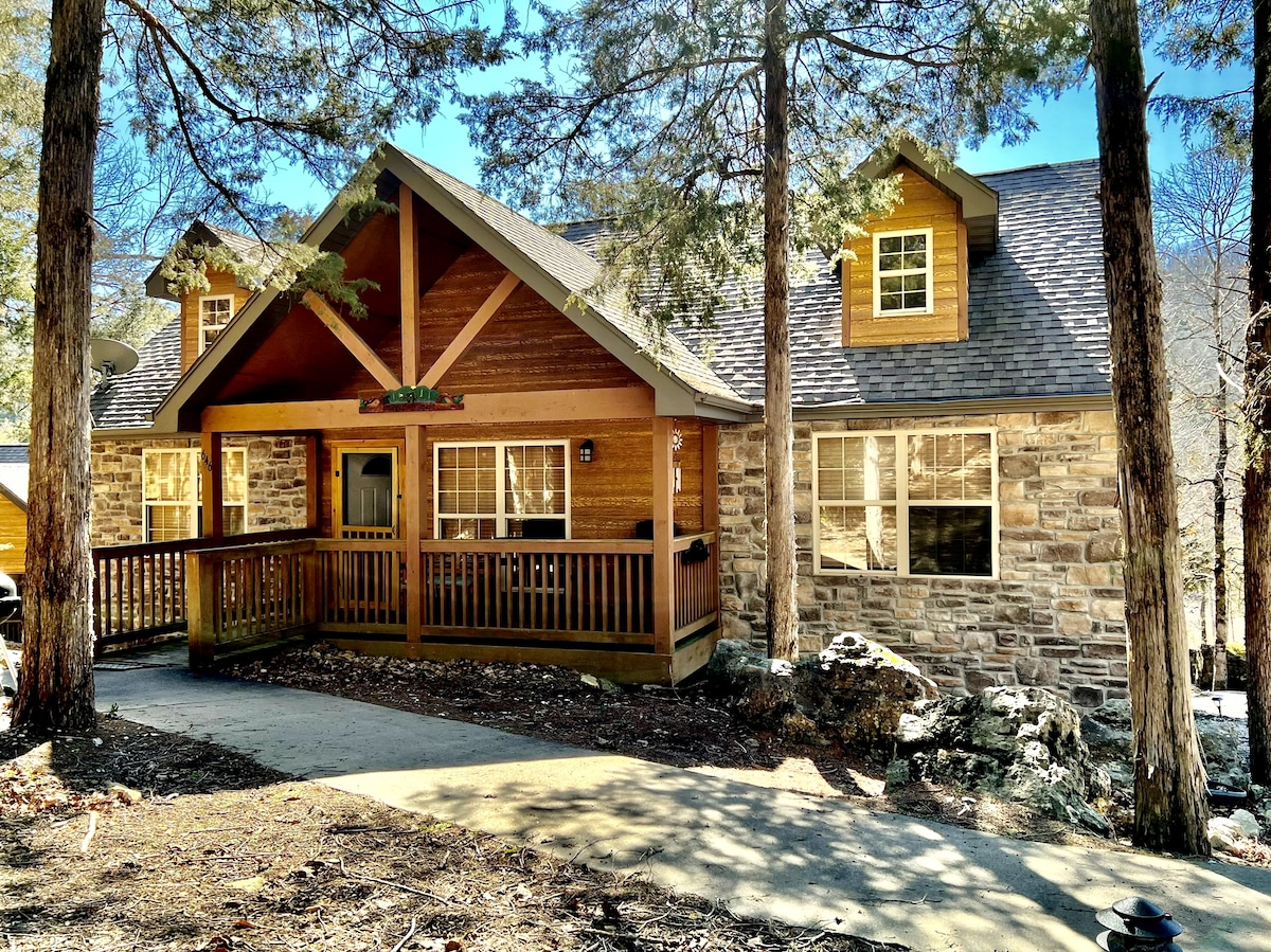 The exterior of the cabin features a rustic design with a mix of wood and stone elements. A spacious front porch is framed by trees, offering a welcoming entrance. Large windows provide natural light, with a clear path leading up to the entrance.
