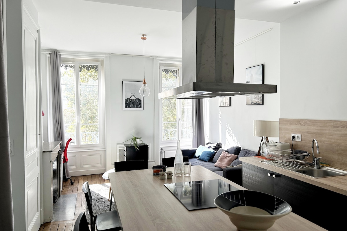 A spacious kitchen area is highlighted by a central island with bar stools and modern appliances. Large windows allow natural light to fill the space, showcasing a cozy living area with a comfortable sofa and a small work desk. Elegant decor complements the inviting atmosphere.