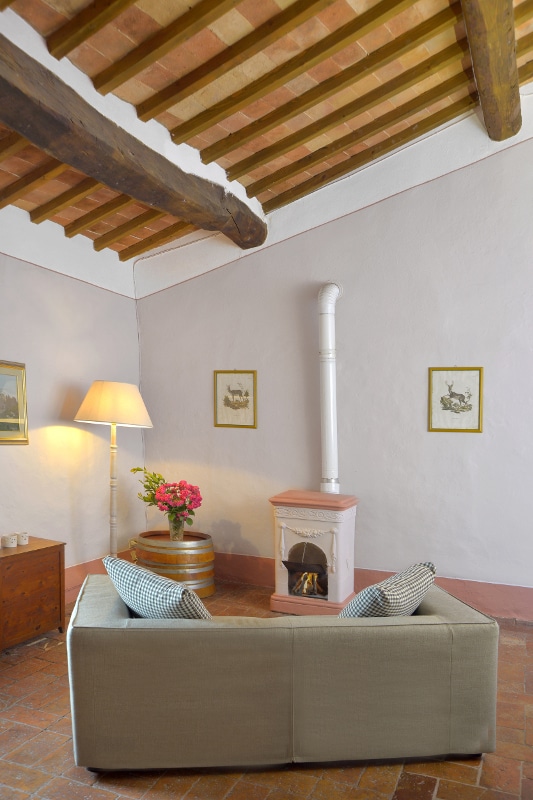 A cozy living area is highlighted by exposed wooden beams and light pink walls. A small terracotta stove stands at one corner, while a simple lamp illuminates the space. Decorative floral arrangements and framed artwork add a touch of character.