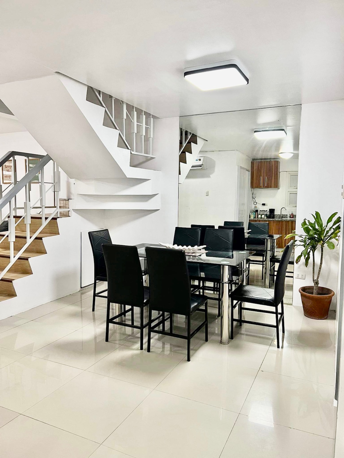 The dining area features a sleek table surrounded by black chairs, creating a modern and inviting setting. Natural light enhances the space, which is complemented by a potted plant and stairs leading to the upper level.